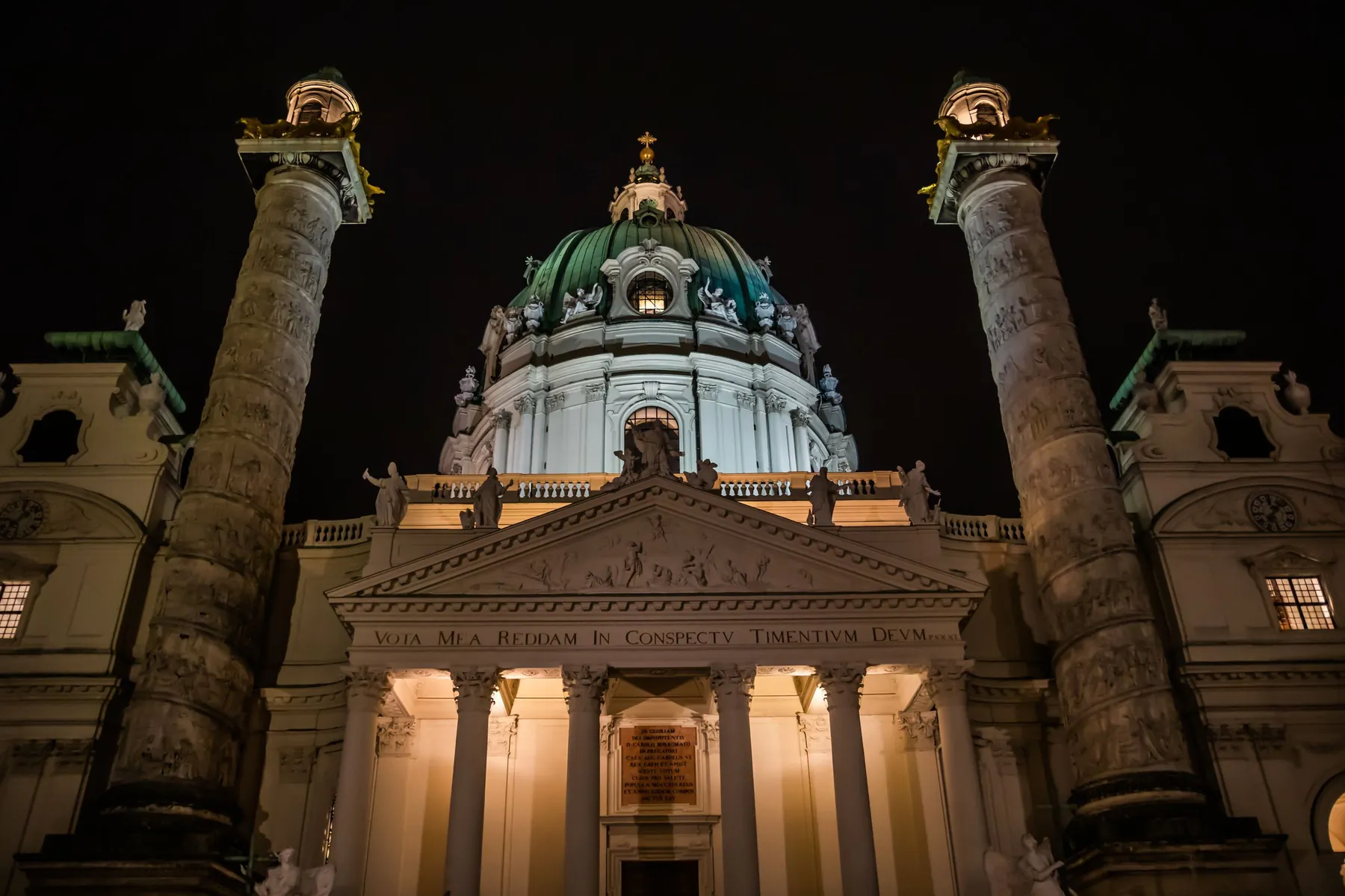 Karlskirche lit up at night, its portico glowing between the two tall columns beneath the green dome